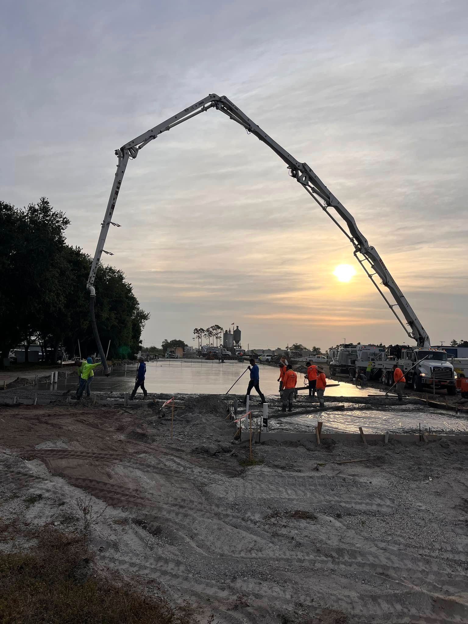 Pump boom arched over a slab pour at sunset, with the crew working the wet concrete and mixer trucks lined up behind