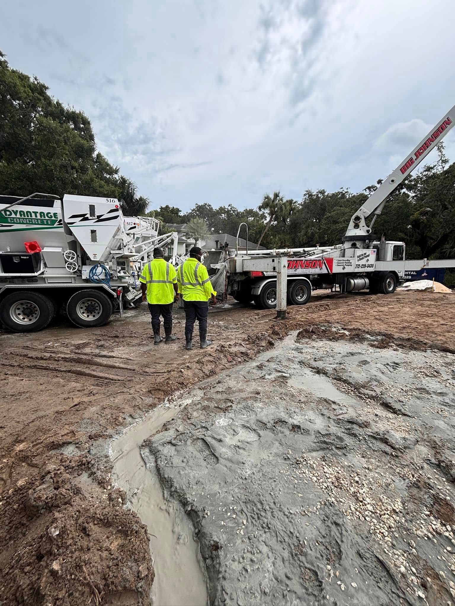 Two crew members in hi-vis shirts walking toward an Advantage Concrete mixer truck, with a pump boom behind and fresh concrete in a trench in the foreground