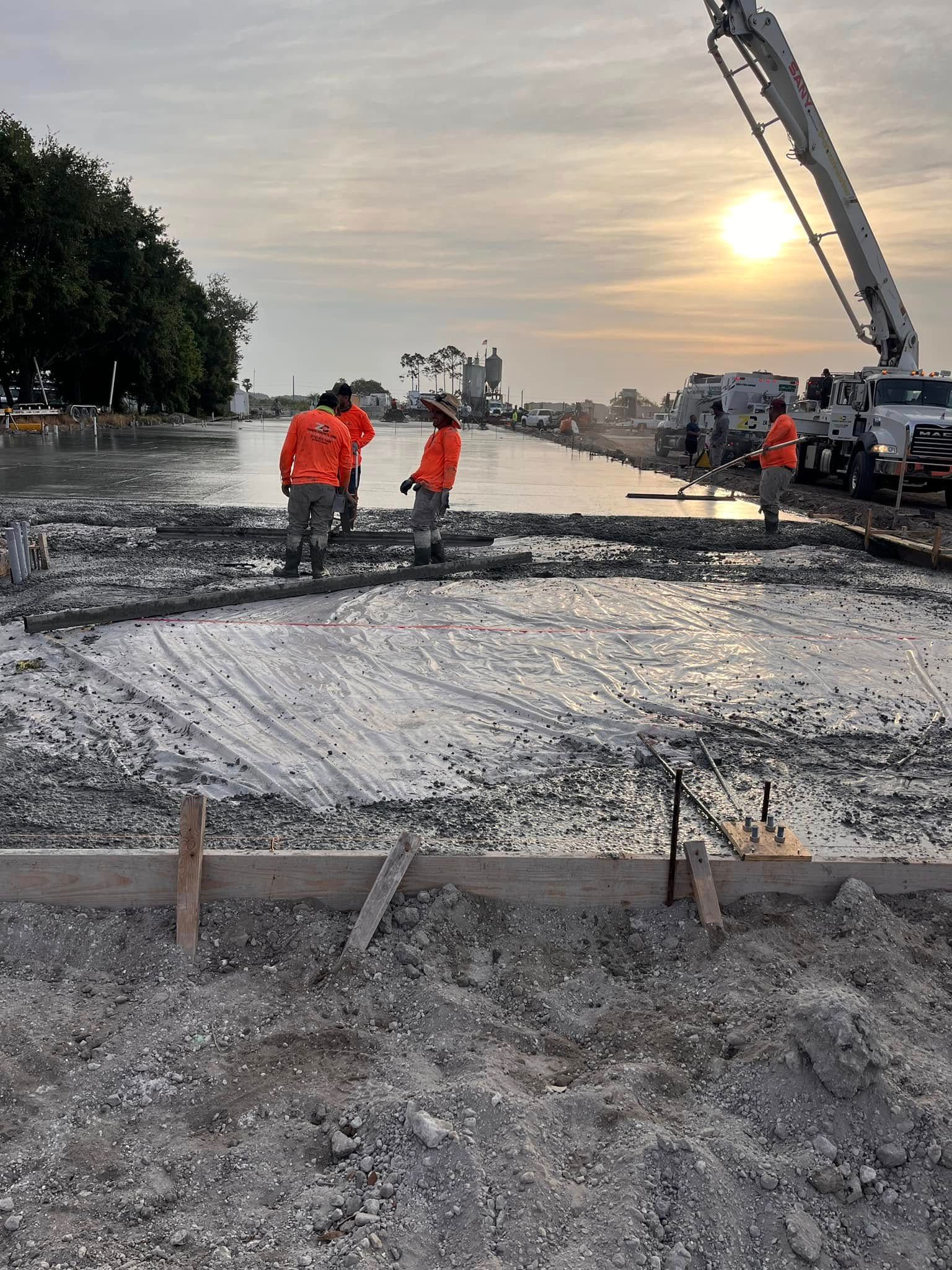 Crew in orange shirts screeding a fresh concrete slab at sunset, with form stakes in the foreground
