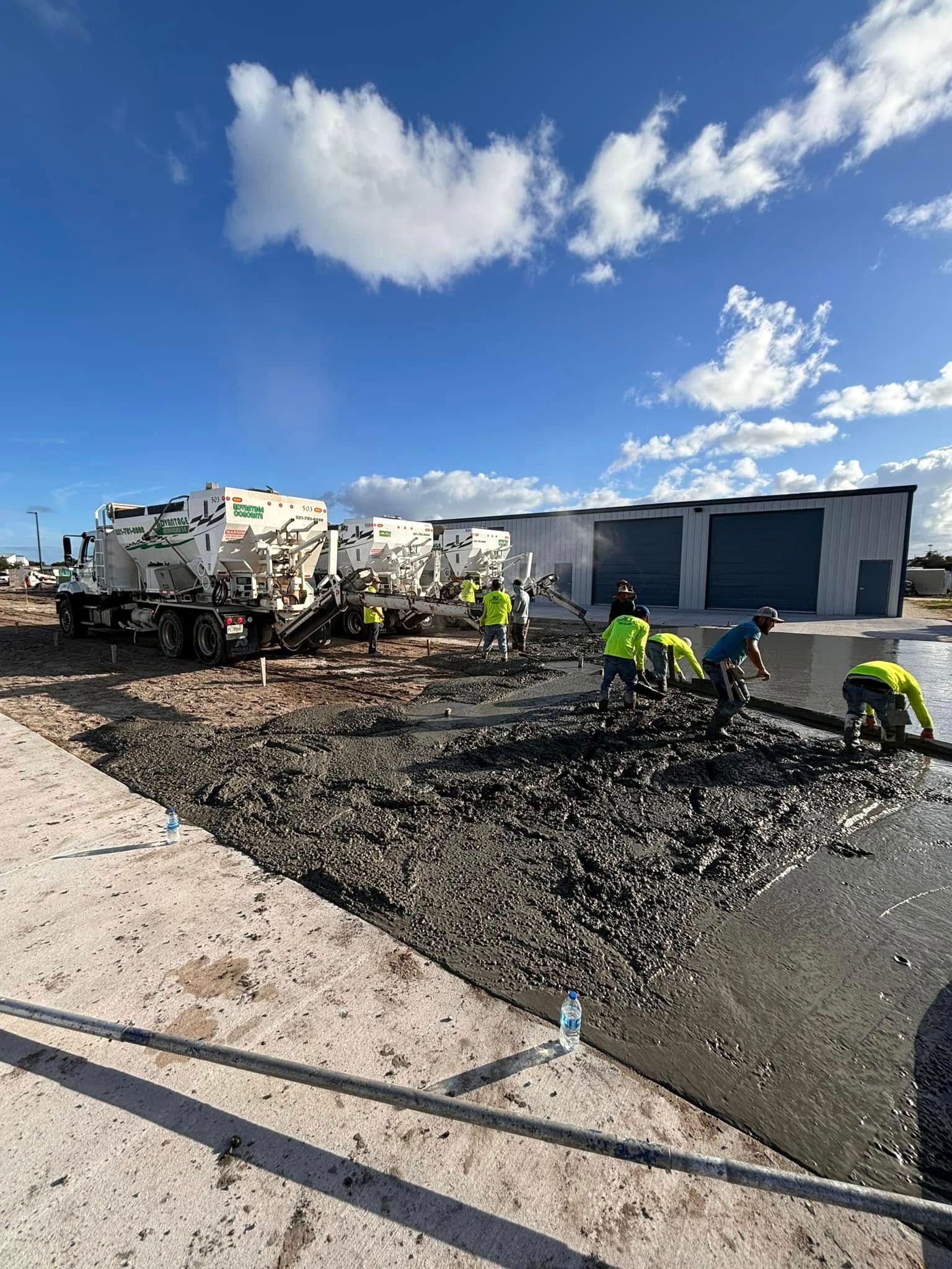 Crew in hi-vis shirts spreading wet concrete on a large slab, with white mixer trucks and a metal building behind