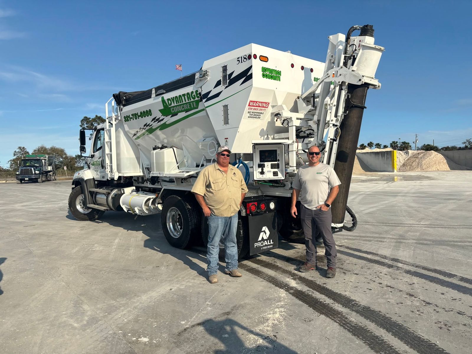 Two crew members in front of a white Advantage Concrete volumetric mixer truck with green lettering, on a paved yard with sand piles and palm trees behind