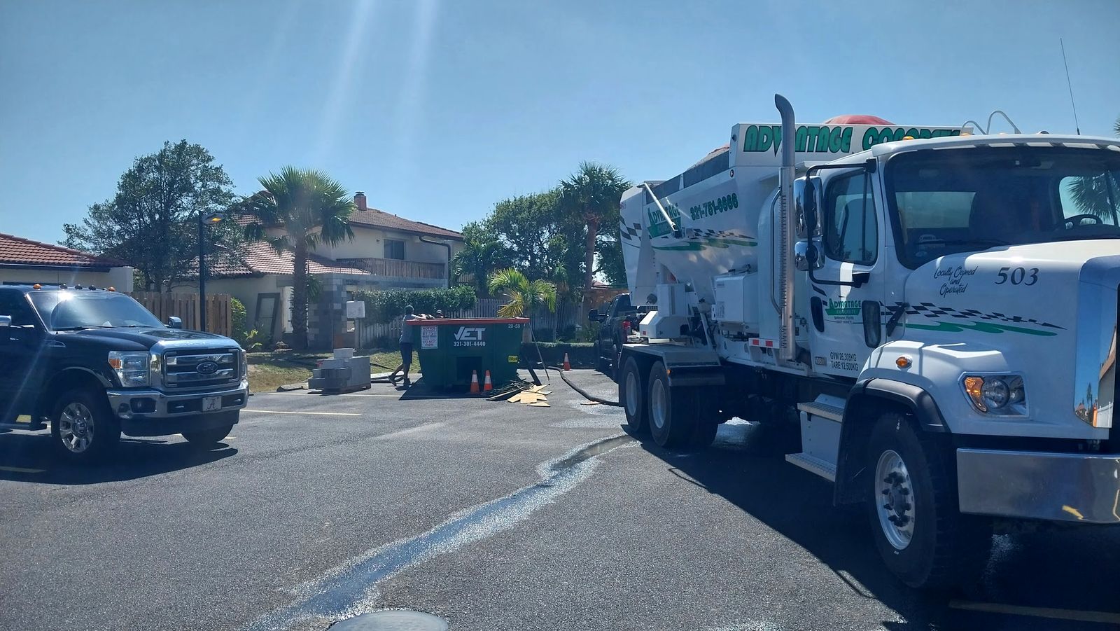 White Advantage Concrete mixer truck parked on a residential street beside a pickup truck, with palm trees and homes behind